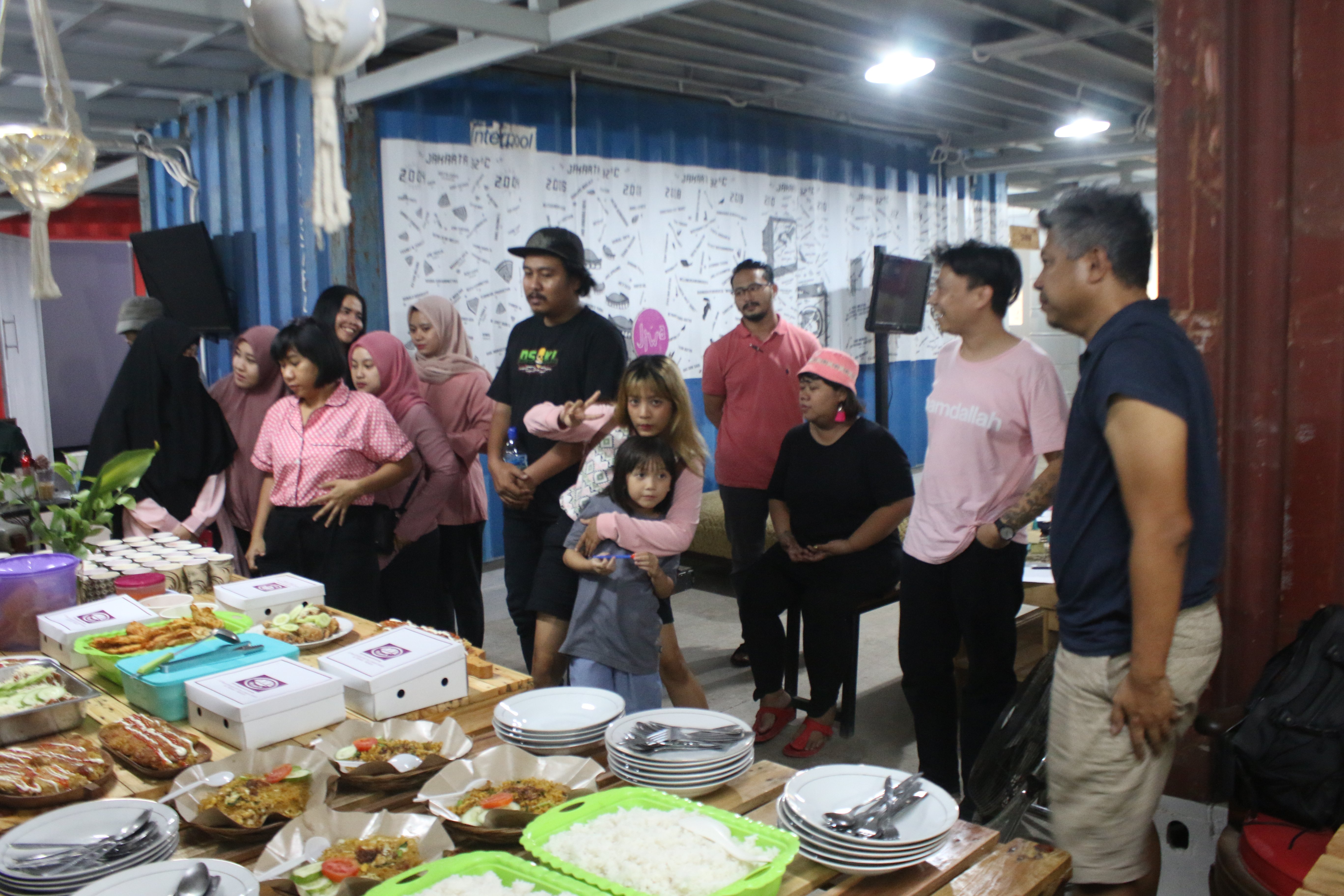 Gudskul members standing around a table filled with food. One person is holding up a v peace sign looking directly at the camera.