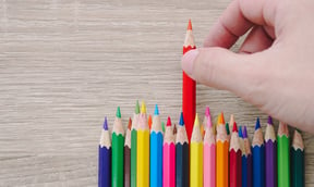 A series of colored pencils stacked next to each other and a hand selecting the red pencil