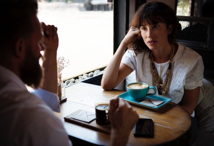 A woman left of frame having coffee at a small wooden table with a bearded man right of frame