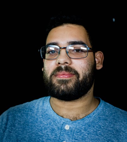 Edwin Torres, Bronx Film 48 Cofounder, with bear and glasses in light blue collarless shirt, standing against black background