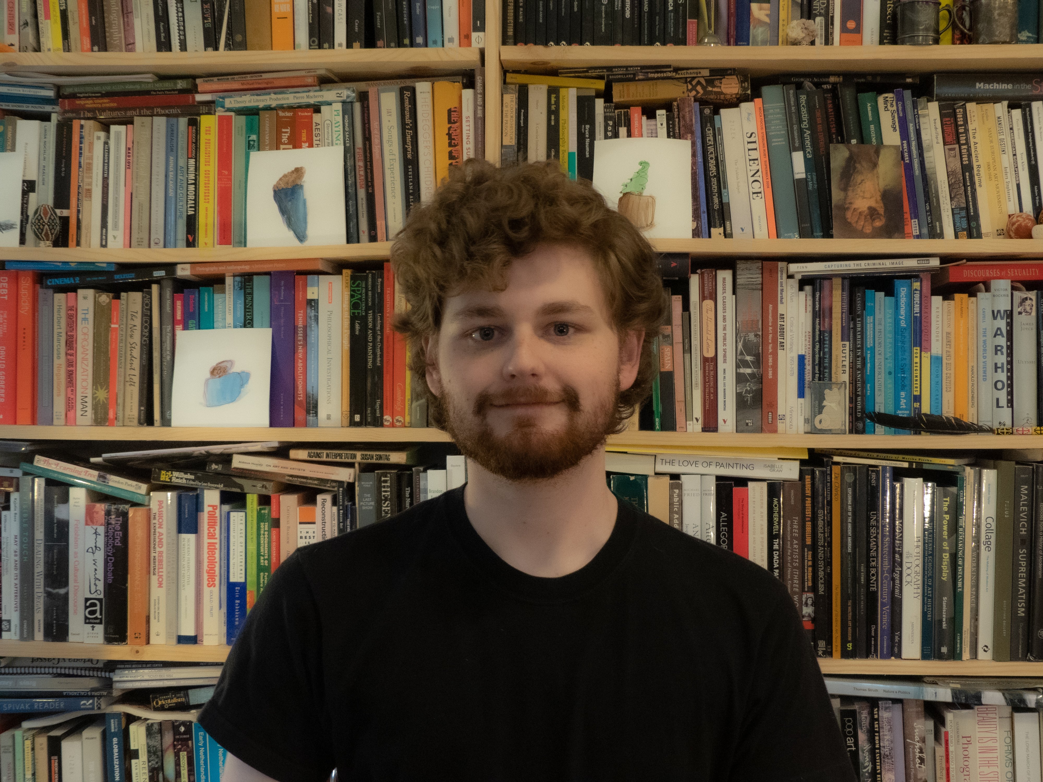 A photo of Colo in front of full bookshelves with full head of hair and beard wearing a black tee