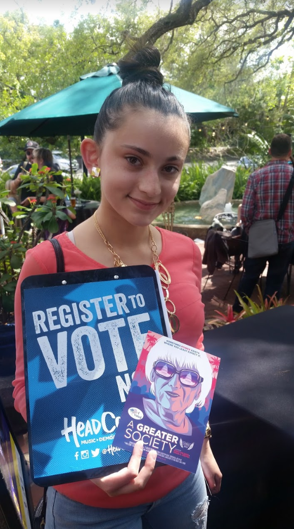 a young person holds the flyer for the movie and a poster encouraging people to register to vote