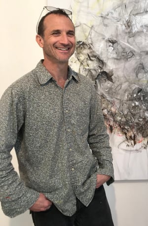 Andrew Purchin in grey shirt with glasses in front of a black and white piece of art