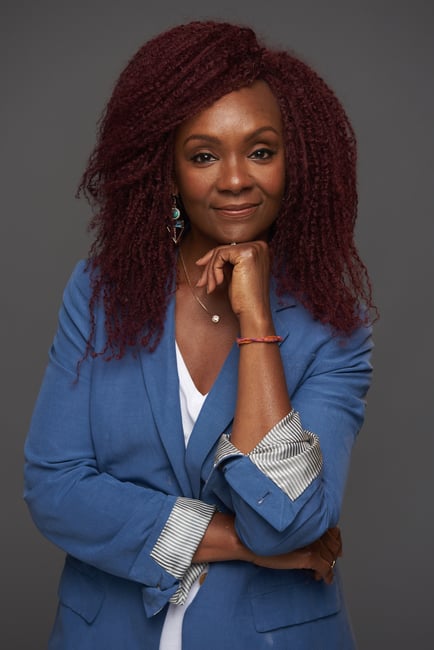 Rha Goddess standing with blue blazer and white shirt with arms folded and hand on chin