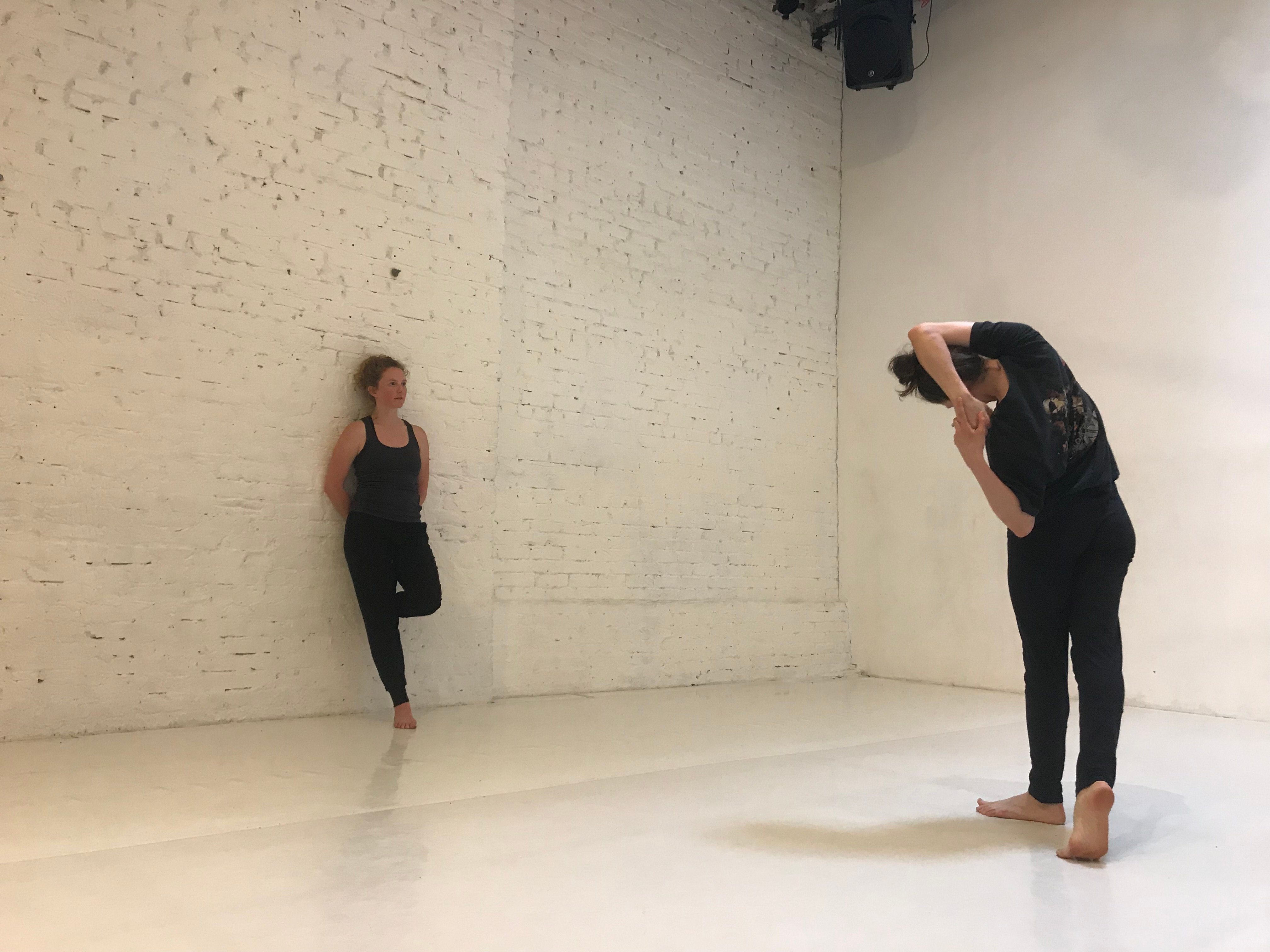 Kelly Todd leans against a wall in a dance studio with another dancer stretching across from her