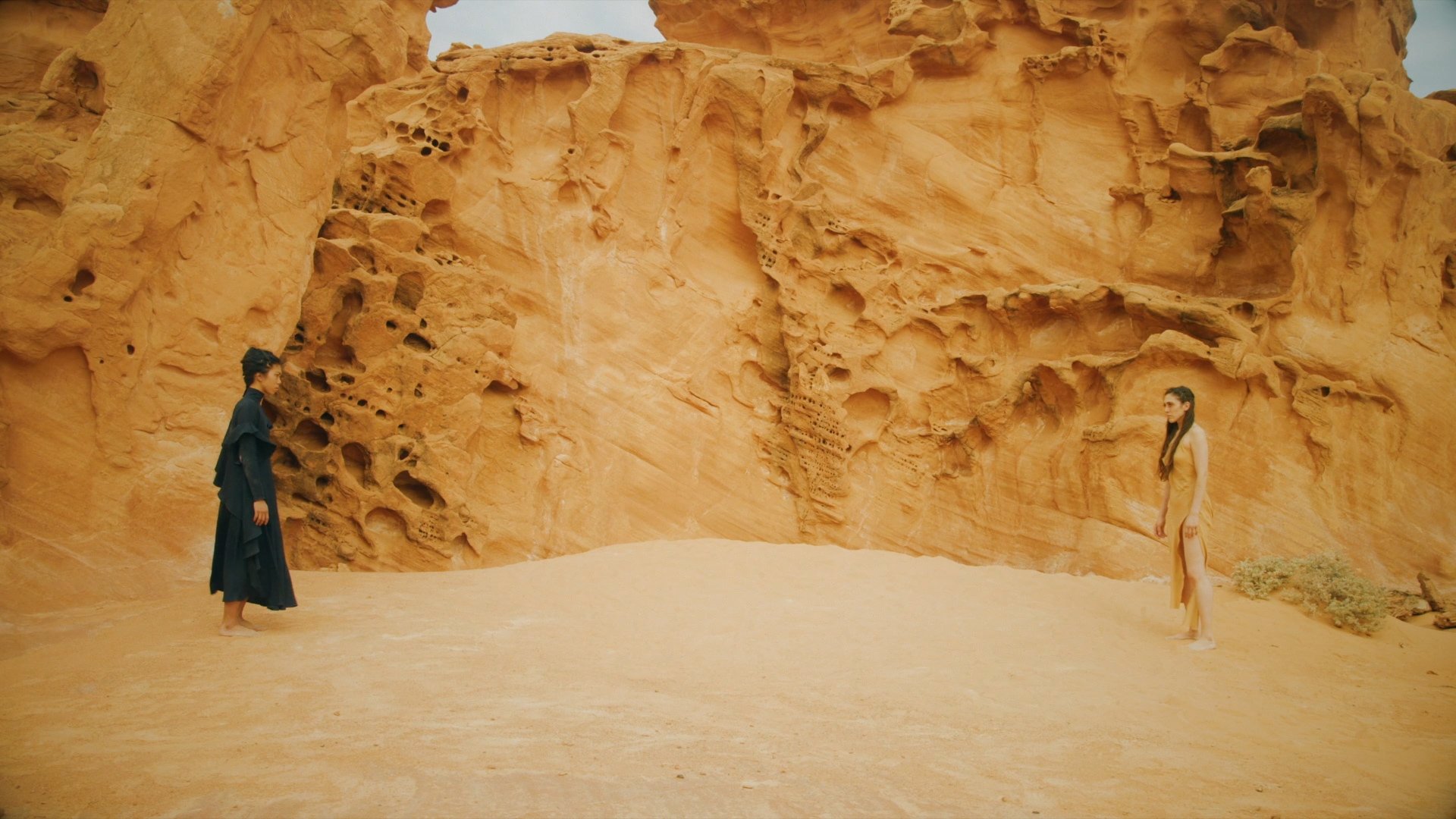 Two people standing across from each other in the desert with a rock formation backdrop