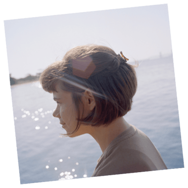 A photo of Laurel Schwulst in front of a large expanse of grey, glistening water. Her hair is tucked behind her ear and she is facing away, looking into the distance with a faint smile. The photos was taken by Jason Fulford, at Plumb Beach in 2021.