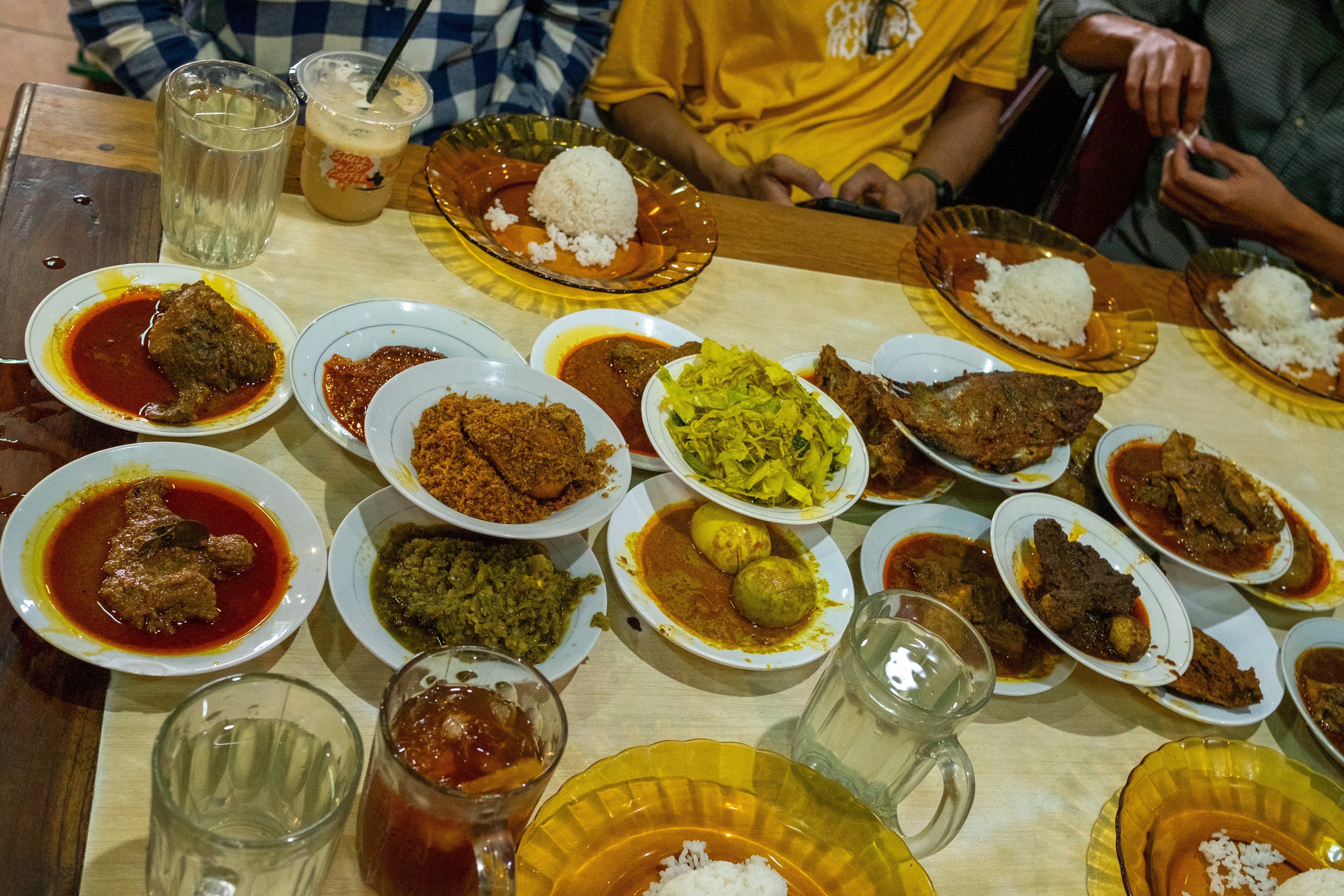 A tan and white table filled with various shared food items. A group of people from lumbung Indonesia's assembly are gathered around it.