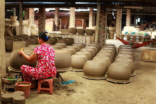 An artisan wearing a red, patterned ensemble works on a large sculpted pot in front of many rows of similar pots.